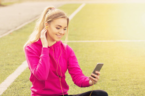 Portrait of young beautiful blonde hair fitness girl using her phone on the stadium. Summer sport activity. Green stadium grass on background. - Photo, image