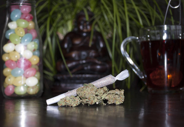 pěkné closeup válcované marihuany plevel kloubu a pupeny, na dřevěné pozadí, soška Buddhy, barevné jellybeans a šálek čaje rooibos - Fotografie, Obrázek