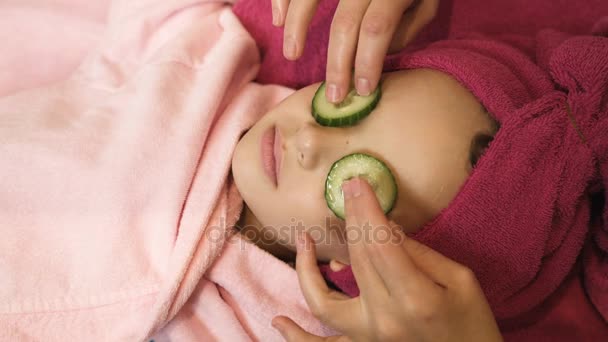 Professional beautician putting cucumber slices on little girls eyes at beauty salon. Spa therapy. Close up view - Footage, Video