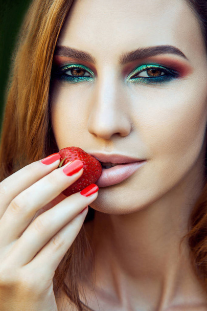 Young beautiful happy funny girl with red dress and makeup holding strawberry in summertime in the park. healthy lifestyle, diet beauty and happiness concept. - Fotó, kép