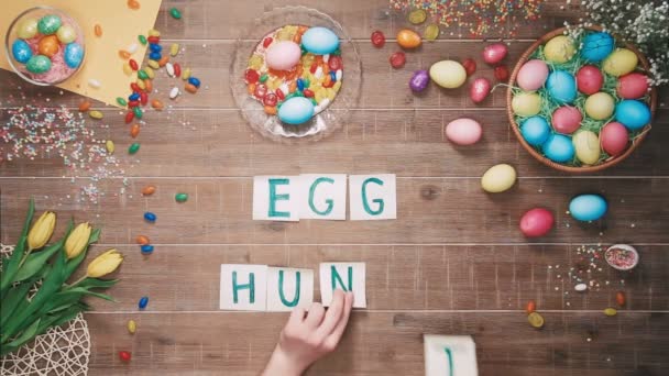 Man puts words Egg hunt on table decorated with easter eggs. Top view - Πλάνα, βίντεο