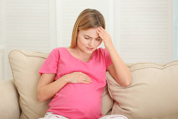 Pregnant woman suffering from headaches while sitting on sofa at home - Фото, изображение