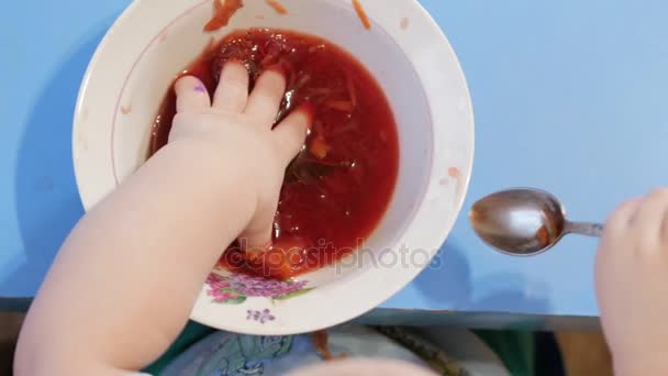An attractive boy 2 years old is eating red soup himself. He climbs his hands in the plate. Table top view. - Imágenes, Vídeo