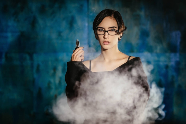 A girl with glasses vaping and releases a cloud of vapor. Model in a black vaper smoke vaporizer on a turquoise background - Foto, Imagem