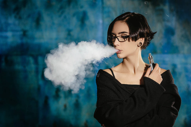 Bir kızla gözlük vaping ve Buhar bulutu serbest bırakır. Turkuaz bir arka plan üzerinde siyah vaper duman Buharlaştırıcı model - Fotoğraf, Görsel