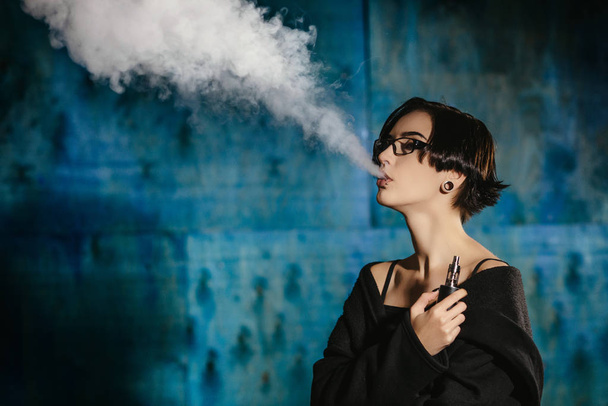 Dziewczyna z vaping okulary i wypuszcza chmurę pary wodnej. Model w internetowych czarny dym odparowalnik na turkus tło - Zdjęcie, obraz