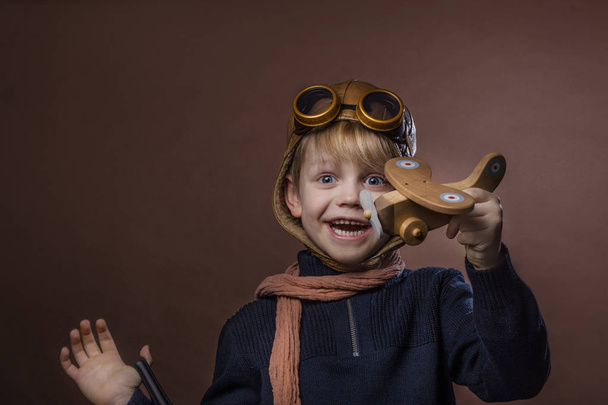 Šťastné dítě oblečené v pilotní čepice a brýle. Kluk hraje s dřevěná hračka letadlo. Koncept sen a svobodu. Retro tónovaný. Studiový portrét nad hnědým pozadím - Fotografie, Obrázek