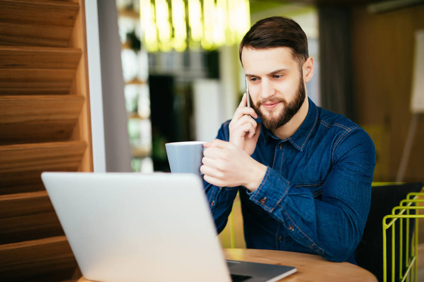 Young handsome man with beard sitting in cafe talking mobile phone, holding cup of coffee and smiling. Laptop on wooden table. - 写真・画像