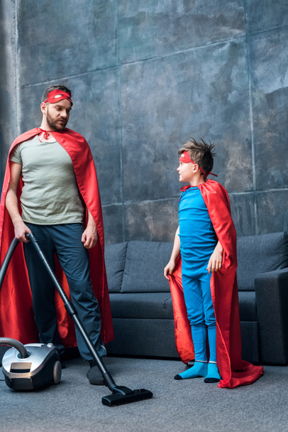 Vater und Sohn in Superheldenkostümen beim Staubsaugen  - Foto, Bild