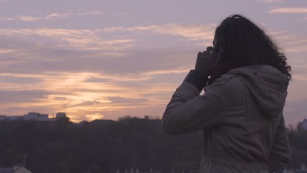 Приваблива змішана раса жінка говорить через смартфон на тлі красивого заходу сонця неба
 - Кадри, відео