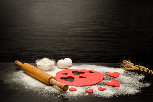 Masa roja con corazones tallados sobre una mesa negra. Rodillo, harina, huevos y espigas de trigo. Lugar para el texto
 - Foto, Imagen