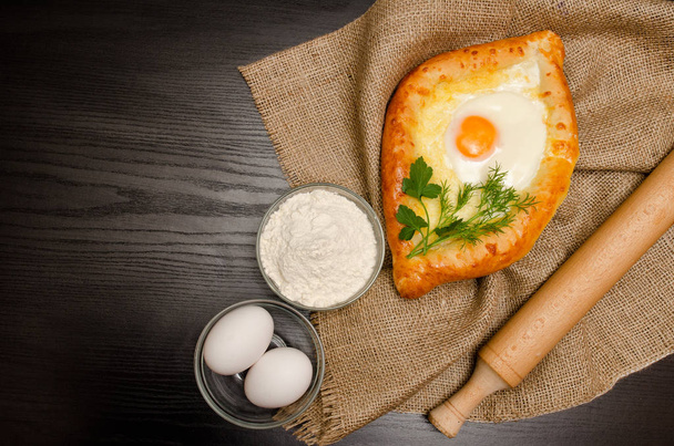 Georgian cuisine. Top view of khachapuri on sackcloth, flour, eggs and rolling pin. Black table, space for text - Foto, Imagen