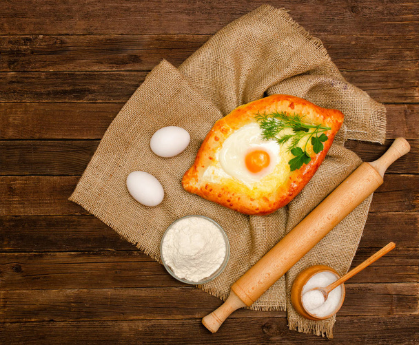 Cuisine géorgienne. Khachapuri aux œufs sur un sac, sel, farine, œufs et persil sur une table en bois
 - Photo, image