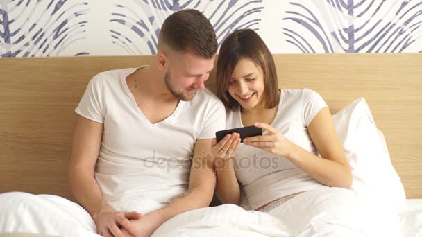 Beautiful young happy couple in bed with mobile phone smiling. Domestic atmosphere, lifestyle photo. - Filmagem, Vídeo