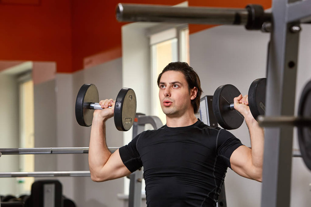 Personal Trainer in black t-shirt doing sitting dumbbell curls for training his biceps, in a gym - Фото, зображення