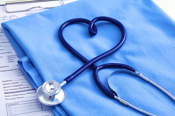 Medische stethoscoop twisted in hart vorm liggen op lijst patiënt medische geschiedenis en blauwe arts uniforme close-up. Medische hulp of verzekering concept. Cardiologie zorg, gezondheid, bescherming en preventie - Foto, afbeelding