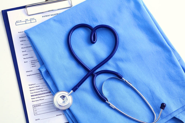 Medical stethoscope twisted in heart shape lying on patient medical history list and blue doctor uniform closeup. Medical help or insurance concept. Cardiology care, health, protection and prevention - Photo, Image