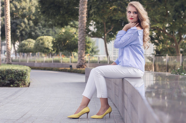 junge Frau mit lockigem blondem Haar sitzt im Park in Dubai  - Foto, Bild