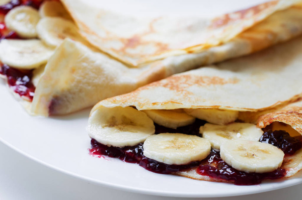 Panqueques finos con mermelada de cereza y rodajas de plátano en un plato blanco
 - Foto, Imagen