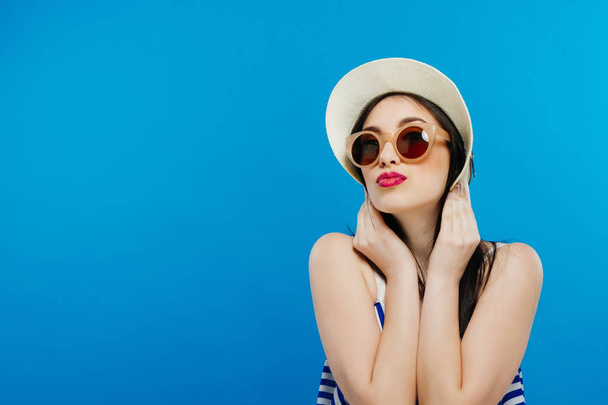 Cheerful beautiful girl in hat and sun glasses. Smiling widely, looking and pointing aside. Summer outfit. Waist up, studio, indoors - Photo, Image