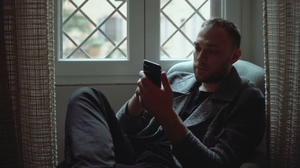 Portrait of attractive man sitting in armchair in living room and using smartphone. Stylish businessman browse Internet. - 写真・画像