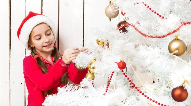 little girl in red santa hat decorating new year tree - Фото, изображение