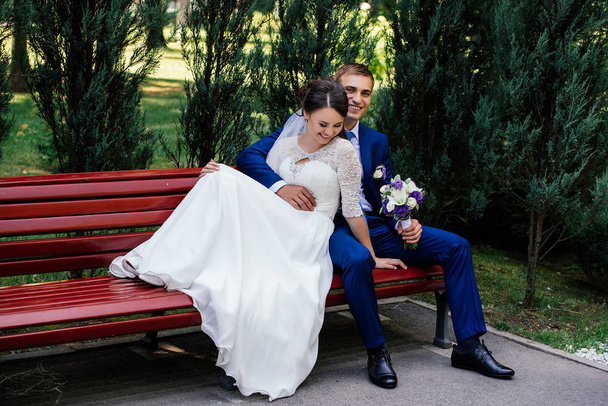 The bride and groom sit on a bench. The groom hugs the bride - Foto, afbeelding