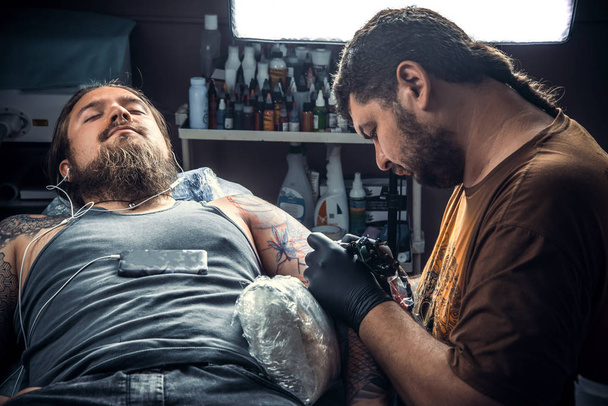 Ammattimainen tatuoija poseeraa tatuointistudiossa. / Master tekee viileän tatuoinnin tatuointistudiossa
. - Valokuva, kuva