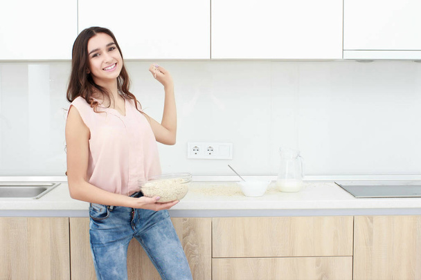 Healthy breakfast . Young beautiful cheerful woman putting cereal in a bowl while sitting on the worktop and smiling. - Fotografie, Obrázek