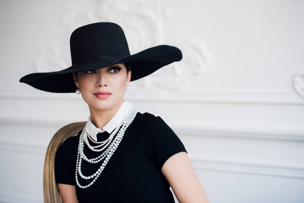 Portret van een mooie jonge vrouw in retro stijl in een elegante zwarte hoed en kleding over luxe rococco muur achtergrond - Foto, afbeelding