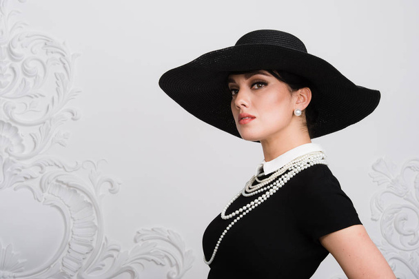 Portrait of a beautiful young woman in retro style in an elegant black hat and dress over luxury rococco wall background - Foto, Imagem
