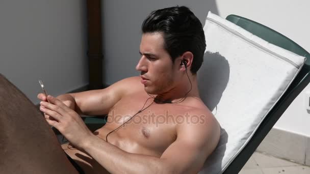Shirtless muscular young man sunbathing, listening to music - Video