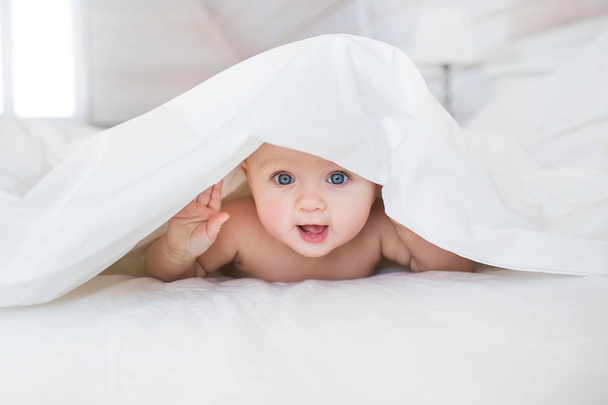 Pretty Infant hides under a white cover in bed in bedroom - Foto, afbeelding