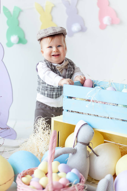 Little boy is dressed in cap. He stands next to box with Easter eggs and hares. he smiles - Photo, Image
