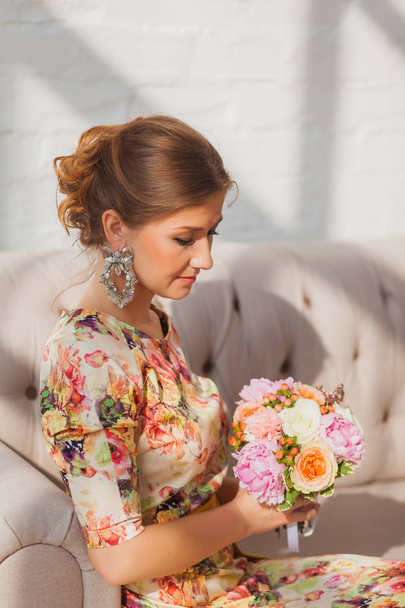 Beautiful girl in a long dress sitting on a sofa with a bouquet - Фото, зображення