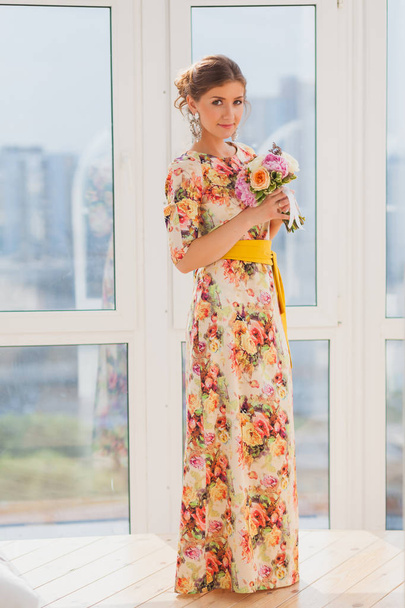 Menina bonita em um vestido longo de pé perto de uma janela com um buquê
 - Foto, Imagem