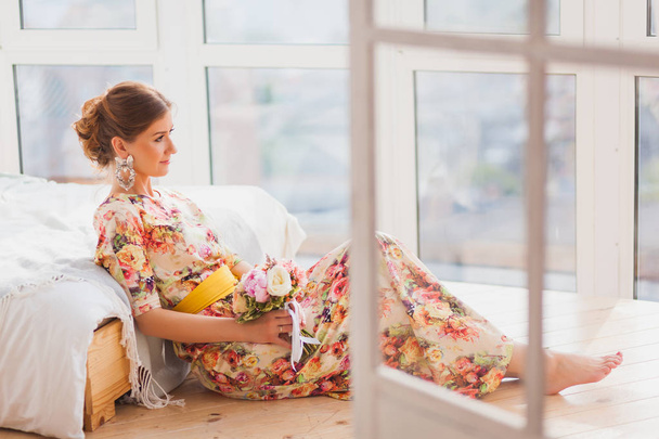 Beautiful girl in a long dress sitting on a sofa with a bouquet - Фото, изображение