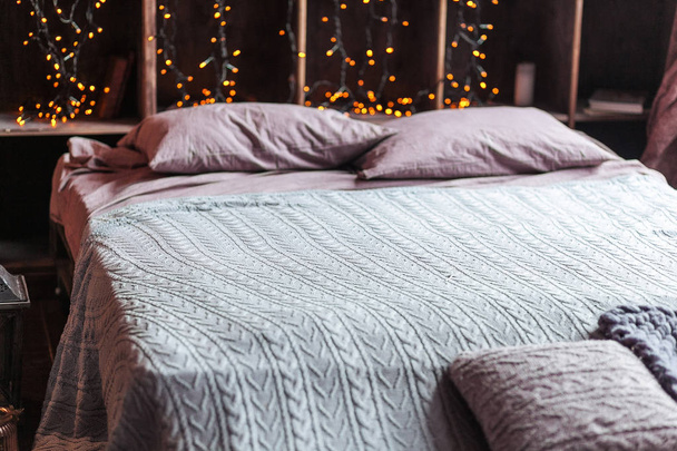 coziness, comfort, interior and holidays concept - cozy bedroom with bed and garland lights at home. A rack with books behind the bed. Candles, a lamp and a lamp stand near the bed. Plaid hand-knitted - Foto, Bild