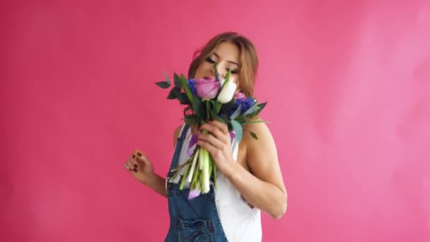 Nice young blonde with blue eyes and a healthy long blond hair smelling flowers on a pink background in jeans - Filmati, video