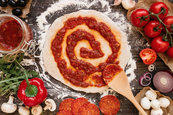 Cooking pizza. Adding fresh tomato sauce to pizza dough. Pizza ingredients on the wooden table top view - Foto, immagini