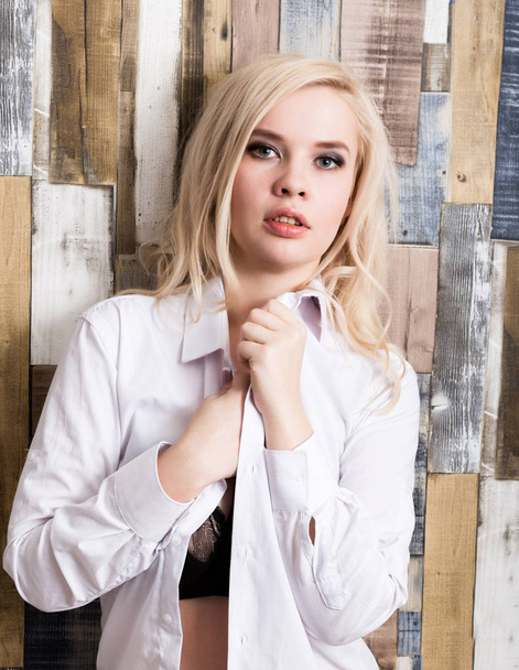 portrait of attractive blonde girl standing on wood wall background. She has blue eyes and dressed in a mans shirt - Foto, Imagen