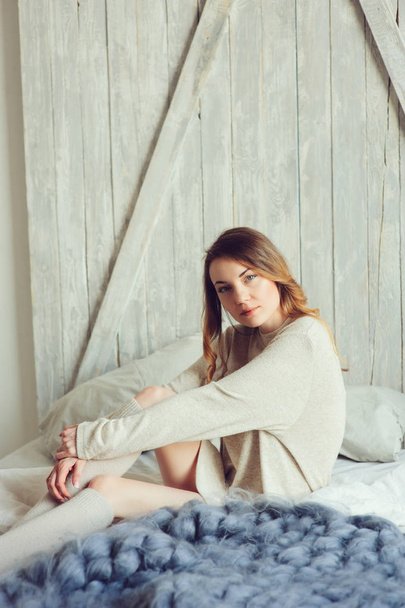 молодая женщина в вязаном кардигане и теплых носках просыпается утром в уютной скандинавской спальне и сидит на кровати с белым постельным бельем. Свободный образ жизни в современном интерьере
 - Фото, изображение