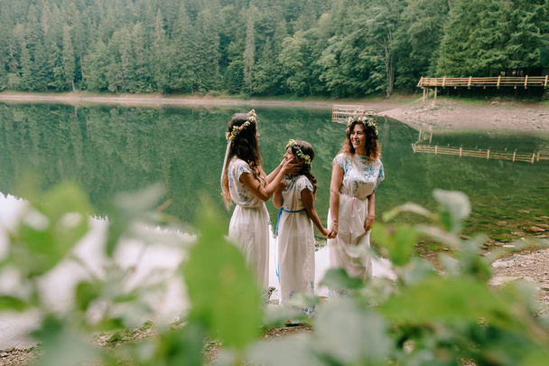 Anne ve iki kızı beyaz elbiseler kır çiçekleri çelenk ile göle yakın yürüyüş - Fotoğraf, Görsel