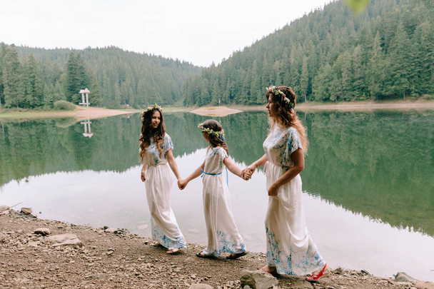 Mãe e duas filhas andando perto do lago em vestidos brancos com grinaldas de flores silvestres
 - Foto, Imagem