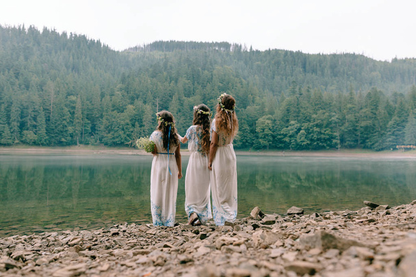 Mom and two daughters walking near the lake in white dresses with wreaths of wildflowers - Photo, Image