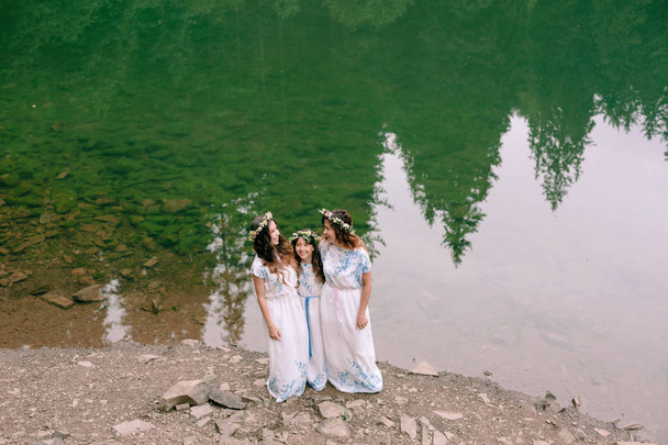 Mama i dwie córki, chodzenie w pobliżu jeziora w białe sukienki z wieńce kwiatów - Zdjęcie, obraz