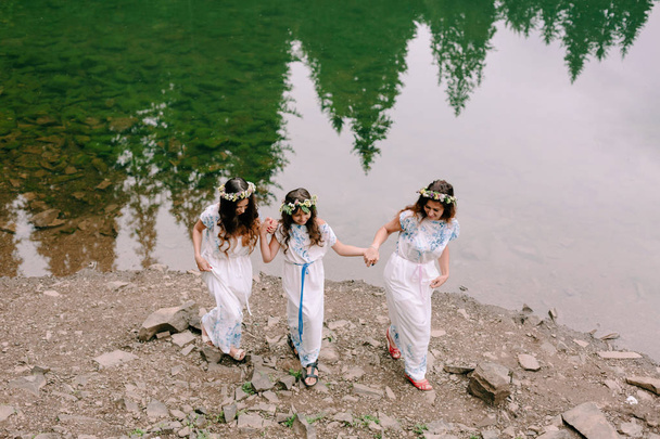 Mamá y dos hijas caminando cerca del lago en vestidos blancos con coronas de flores silvestres
 - Foto, imagen