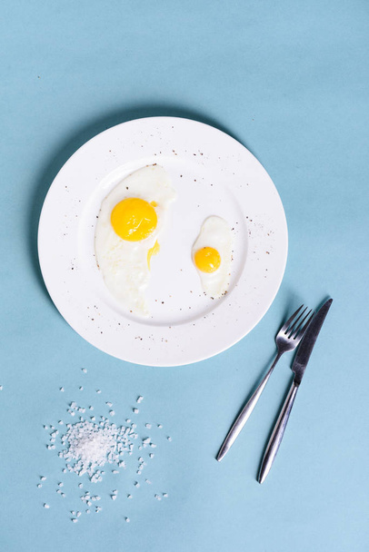 Healthy delicious breakfast of fried eggs with salt and pepper in a white plate on a blue table background. Top view, text space, - 写真・画像