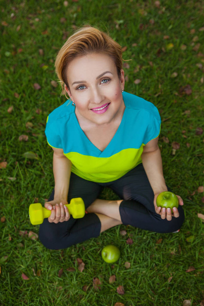 Sporty woman with dumbbells is sitting on the green grass outdoor. - Fotó, kép