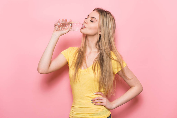 Beautiful young fitness woman with perfect body shape drinking fresh water from plastic bottle standing near pink wall. - Fotoğraf, Görsel